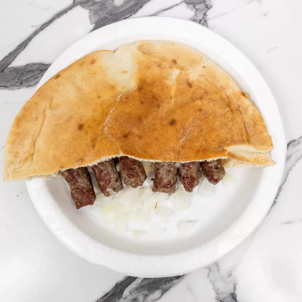 Cevapi in lepinja bread with diced onions on a white paper plate.