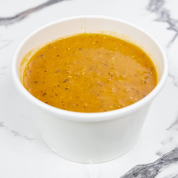 A white paper cup filled with a rich orange-brown soup, speckled with herbs, on a marble surface.