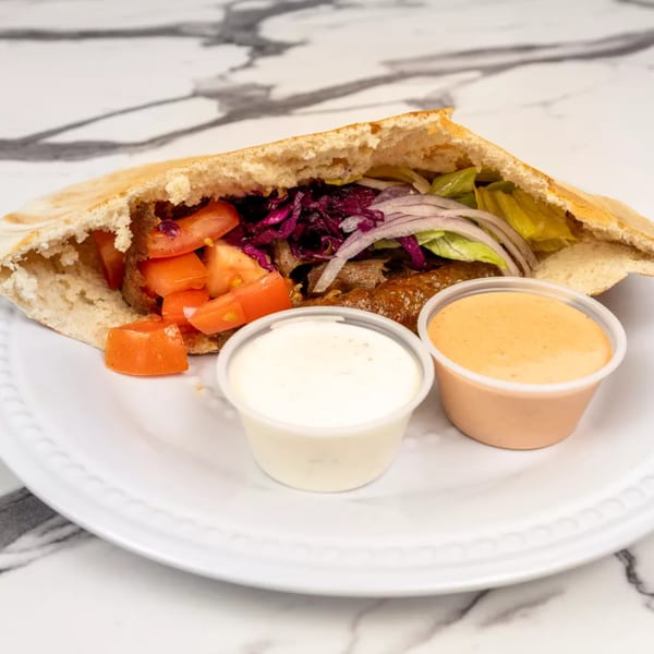Pita bread sandwich filled with meat, tomatoes, red cabbage, onions, and lettuce, with two sauce cups.