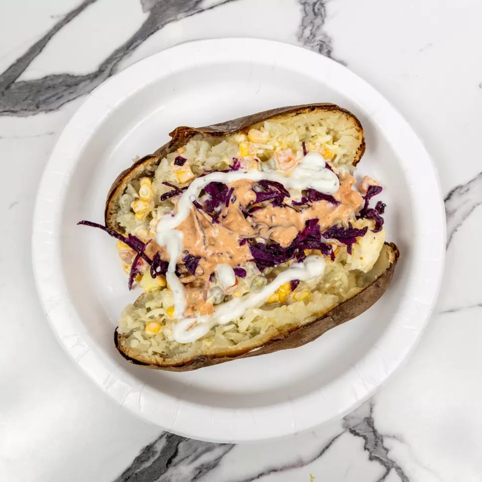 A baked potato stuffed with corn, purple cabbage, and drizzled with white and orange sauces on a white plate.