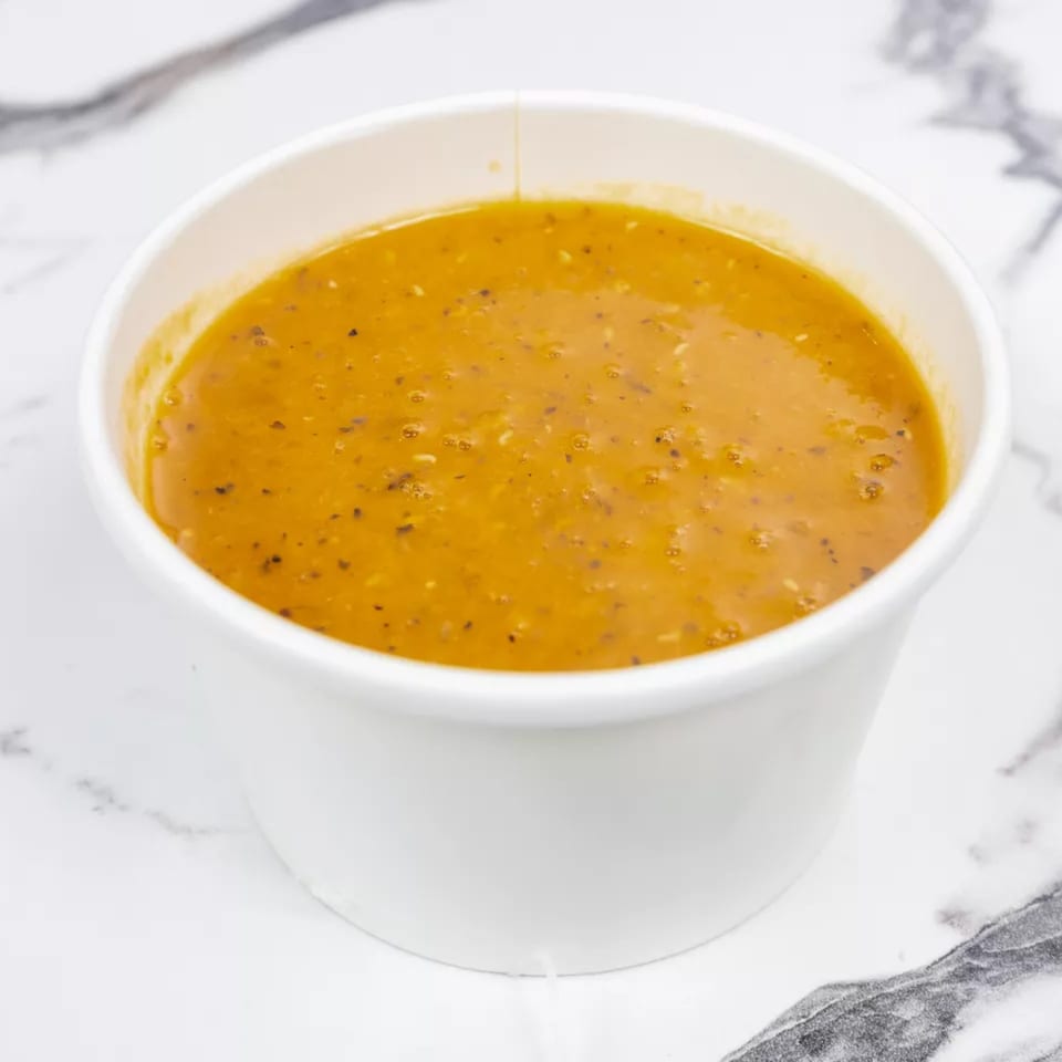 A white paper cup filled with a rich orange-brown soup, speckled with herbs, on a marble surface.