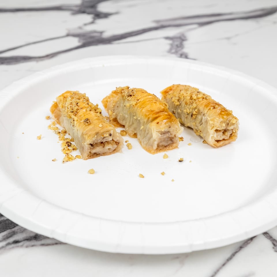 Three golden-brown baklava rolls, topped with chopped nuts, on a white paper plate.