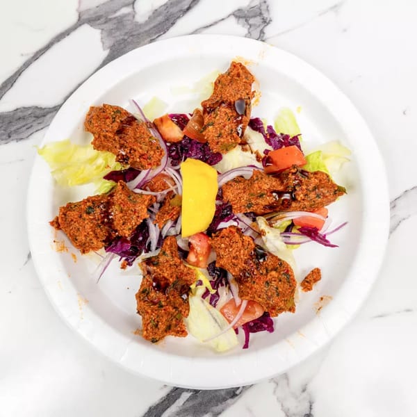 A white plate of çiğ köfte, salad, red cabbage, onions, tomatoes, and a lemon wedge.